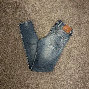 Replay Anbass blå jeans slim fit 29/32 - Snygga Replay Anbass jeans i blå tvätt med slitningar och detaljerade sömmar. Modellen är slim fit med raka ben och klassisk femficksdesign. Jeansen har mellanblå färgton och är gjorda i mjukt bomullsmaterial med lite stretch för extra komfort.