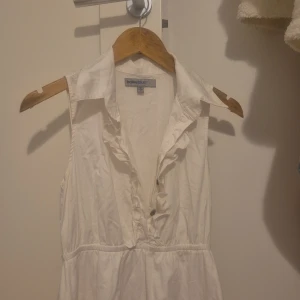 Vit ärmlös klänning från Bailey Blue - Söt vit klänning från Bailey Blue med volangdetaljer längs v-ringningen och krage. Klänningen är ärmlös, har markerad midja och lätt utställd kjol. Perfekt för varma dagar och enkel att styla med accessoarer.