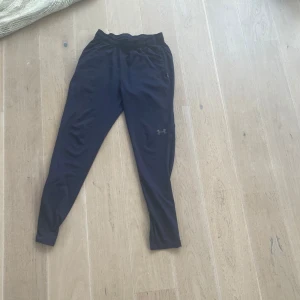 Svarta träningsbyxor Under Armour - Svarta träningsbyxor från Under Armour med smal passform och elastisk midja. Byxorna har diskret logga på benet och är gjorda i ett lätt och stretchigt material som passar perfekt för träning eller chill.