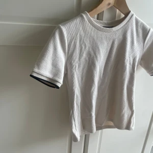 Vit ribbad t-shirt från Zara - Vit ribbad t-shirt från Zara med korta ärmar och rund hals. Ärmsluten har en svart och glittrig detalj som ger en snygg kontrast. T-shirten är i ett mjukt och stretchigt bomullsmaterial, perfekt för en clean och stilren look.
