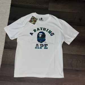 BAPE t-shirt - Helt ny Vit t-shirt från BAPE med klassisk 'A Bathing Ape'-logga framtill i blågrön camo-stil. Rund halsringning och korta ärmar. Materialet är mjuk bomull och passformen är normal. Perfekt för dig som gillar streetwear och vill sticka ut. Jag på bilden är ca 182cm och väger 82kg