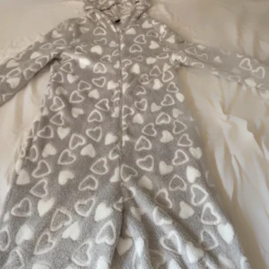 Grå fleecehoodie med hjärtan och öron - Supermysig grå hoodie i fleece med vita hjärtan över hela och gulliga öron på huvan. Långärmad och hel dragkedja framtill, perfekt för chilliga dagar hemma. Oversized känsla och riktigt fluffig, passar dig som älskar söta detaljer och komfort.