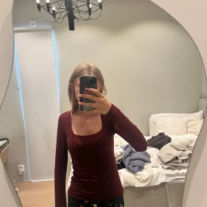 Vinröd långärmad topp med fyrkantig ringning - Vinröd långärmad topp med snygg fyrkantig ringning. Modellen är tight och sitter slimmat mot kroppen. Perfekt till jeans eller kjol för en clean look. Materialet känns mjukt och stretchigt, troligen bomull eller polyesterblandning.