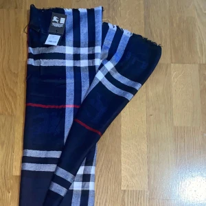 Burberry Scarf - Snygg halsduk från Burberry i klassiskt rutigt mönster. Färgerna är marinblå, vit, svart och en tunn röd linje som ger extra edge. Halsduken är tunn och mjuk, perfekt att slänga över axlarna för en stilren look.