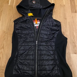 Svart dunväst från Parajumpers - Snygg svart dunväst från Parajumpers med huva och klassisk gul detalj vid halsen. Västen har quiltad framsida, dragkedja hela vägen och sidopaneler i stretchigt material. Perfekt lager-på-lager-plagg med sportig vibe och praktiska fickor. Vill jöttegjärba bara få den såld.