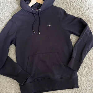 Mörkblå hoodie från GANT XS - Säljer en mörkblå hoodie från GANT i storlek XS. Tröjan har huva med snörning, magficka och diskret GANT-logga broderad på bröstet. Tillverkad i mjuk bomullsmix, perfekt för chill dagar eller när du vill ha en clean och enkel look.