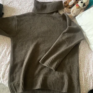 Brun/ grå  polotröja i ull/kashmir från H&M - Mysig grå polotröja från H&M i en blandning av ull och kashmir. Tröjan har ribbade muddar, hög polokrage och lång ärm. Perfekt för kyliga dagar och enkel att styla med jeans eller kjol.