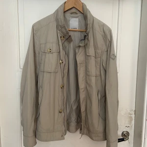 Beige vindjacka från J.Lindeberg - Eftertraktad beige field jacka från J.Lindeberg med klassisk krage, flera fickor framtill och diskret logga på ärmen. Perfekt för sommar och höst när du vill ha en clean och elegant look. Jackan är slutsåld men kostar runt 3000kr ny👊