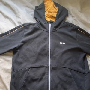 Svart hoodiejacka från BOSS med dragkedja - Svart hoodiejacka från BOSS med gul insida på huvan och vita detaljer. Jackan har dragkedja framtill, fickor och BOSS-logga på bröstet samt längs ärmarna. Snygg kontrast med gul insida och stilrena linjer. Perfekt för en chill och sportig look.