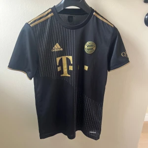 Svart FC Bayern München tröja Adidas - Snygg svart FC Bayern München fotbollströja från Adidas med guldiga detaljer och loggor. Tröjan har korta ärmar, tunna vita prickar framtill och klassiska Adidas-ränder på axlarna. Tillverkad i lätt och ventilerande material, perfekt för match eller träning.
