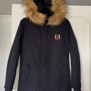 Svart parkasjacka från Svea med fuskpäls - Svart parkasjacka från Equestrian by Svea med avtagbar fuskpäls runt kragen. Jackan har Svea-logga på bröstet, två snedställda fickor och quiltat foder. Perfekt för kalla dagar med sin långa modell och varma känsla. Dragsko i midjan för bättre passform. Väldigt lite använd! 