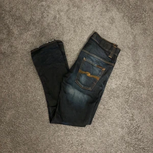 Mörkblå jeans från Nudie Jeans, 30/32 - Säljer ett par mörkblå jeans från Nudie Jeans med snyggt slitna detaljer och klassiska orange sömmar. Modellen har raka ben och fem fickor, samt Nudie-loggan broderad på bakfickorna. Jeansen är i bomull och har en stilren, tidlös look.