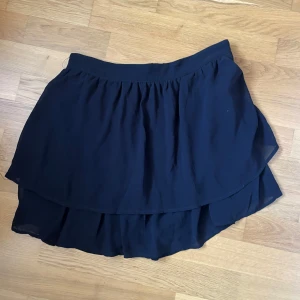 Mörkblå volangkjol i chiffong - Supersöt mörkblå kortkjol med dubbla volanger och flowig känsla. Perfekt för dig som gillar en luftig och lätt stil. Kjolen har resår i midjan och är gjord i ett tunt, svepande material som ger snygg rörelse. Skriv för fler bilder