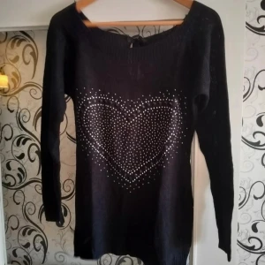Svart stickad tröja med hjärtmönster - Svart stickad tröja med långa ärmar och ett stort hjärta av silvriga nitar framtill. Tröjan har en rund halsringning och är lite längre i modellen, perfekt att matcha med jeans eller leggings.