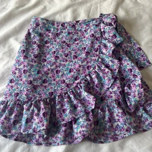 Blommig lila/blå volangkjol  - Supersöt kort kjol med blommigt mönster i lila, blått och grönt. Kjolen har volangkant och en rosett i sidan. Köpt från New Yorker och kommer inte mer till användning. Kontakta gärna innan köp!