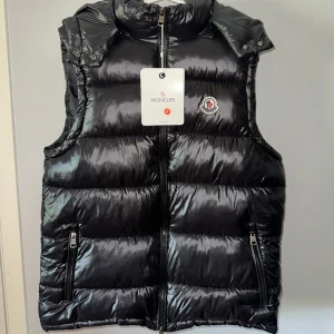 Svart dunväst från Moncler - Snygg svart dunväst från Moncler med glansig finish och klassisk logga på bröstet. Västen har huva, två dragkedjefickor och hel dragkedja framtill. Jag säljer den då min kille fick den som en present och det var inte hans stil, först till kvarn☺️Kan inte så mycket om detta märket så priset kan diskuteras jag lägger pris efter vad jag värderar den.