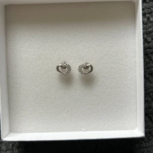 Söta hjärtformade örhängen i silver - Ett par små örhängen i form av hjärtan, tillverkade i silverfärgat material och dekorerade med gnistrande stenar.