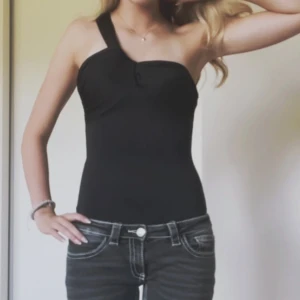 Svart one shoulder body - Snygg svart body med one shoulder-design och smal passform. Toppen är ärmlös och har en bred axelrem på ena sidan för en cool asymmetrisk look. Perfekt att styla med jeans eller kjol för en trendig outfit.