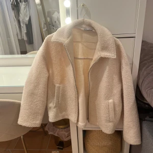 Beige teddyjacka med dragkedja - Supermjuk och fluffig beige teddyjacka med klassisk krage och dragkedja framtill. Jackan har två fickor på sidorna och är perfekt för att hålla värmen under kyliga dagar. Enkel och stilren design som passar till det mesta.