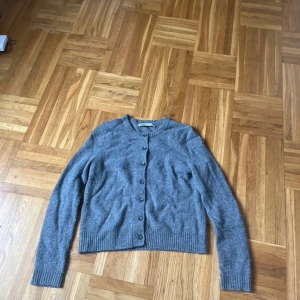 Grå kofta i ull med knappar - Mysig grå kofta i mjuk ull med rund halsringning och knäppning framtill. Långärmad modell med ribbade muddar vid ärmslut och nederkant. Perfekt att slänga över en t-shirt eller ha som lager under jackan.