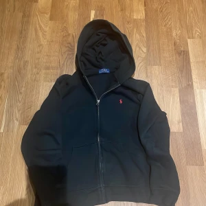 Svart hoodie från Polo Ralph Lauren - Svart hoodie från Polo Ralph Lauren med dragkedja och huva. Klassisk röd broderad logga på bröstet. Mjuk insida och känguruficka framtill. Perfekt för chill dagar eller när du vill ha en clean streetstyle.