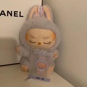 Labubu - Supersöt liten väska från Pop Mart The Monsters, formad som en kanin med stängda ögon och lurvig lila plysch. Väskan har en karbinhake upptill så du kan fästa den på din väska eller nyckelknippa. Perfekt accessoar för dig som gillar gulliga detaljer.