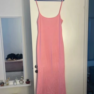 Lång rosa klänning med smala axelband - Enkel och stilren långklänning i rosa med smala axelband och rak passform. Klänningen har en mjuk, slät yta och är perfekt för varma dagar. Passar dig som gillar minimalistisk och feminin stil.