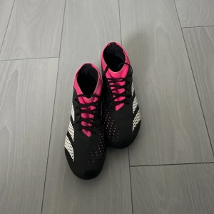 Adidas Predator fotbollsskor svart/rosa - Säljer ett par Adidas Predator fotbollsskor i svart med vita detaljer och rosa inslag. Skorna har snörning och är designade för att ge bra grepp på planen. Snygg och sportig look med klassiska Adidas-ränder på sidorna och rosa mesh runt öppningen.