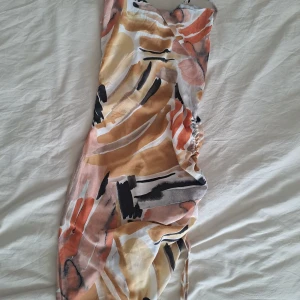 Färgglad kortklänning med snörning - Säljer en kort klänning med abstrakt mönster i beige, orange, svart och vitt. Klänningen har smala axelband, snörning i sidan och en lätt rynkad detalj. Perfekt för sommarens varma dagar och har en flowy känsla.