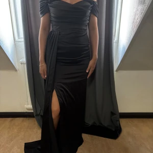 Svart långklänning med slits - Elegant svart långklänning i glansigt material med offshoulder och draperad överdel. Klänningen har en hög slits framtill och figurnära passform som framhäver siluetten. Perfekt för bal eller festliga tillfällen.