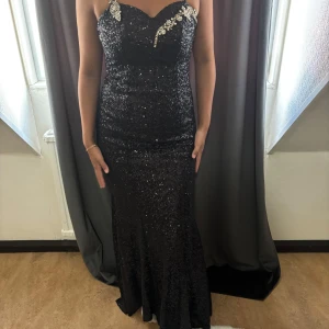 Svart glittrig långklänning med paljetter - Supersnygg svart långklänning med smala axelband och massor av glittrande paljetter. Klänningen har figurnära passform och v-ringad urringning med dekorativa pärldetaljer upptill. Perfekt för bal eller fest där du vill sticka ut.