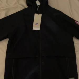 Svart vindjacka från Canada Goose L - Svart vindjacka från Canada Goose med huva och dragkedja framtill. Klassisk logga på ärmen och stilren design. Jackan är i lätt material, perfekt för blåsiga dagar. Snygg och enkel att matcha med allt. Storlek L