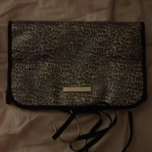 Leopardmönstrad necessär Victoria's Secret - Necessär från Victoria's Secret med leopardmönster i svart och grått. Flera fack på insidan i rosa, perfekt för smink eller småprylar. Praktisk krok för upphängning och knytband för stängning. Snygg och smidig att ta med på resan.