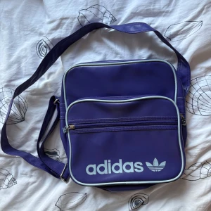 Lila axelväska från Adidas - Retroinspirerad lila axelväska från Adidas med justerbar rem, stora fack och dragkedjor med Adidas-logga. Väskan har ljusblå kantband och ett stort tryckt Adidas-tryck framtill. Perfekt för att bära med sig allt man behöver till skolan eller stan. Oanvänd.