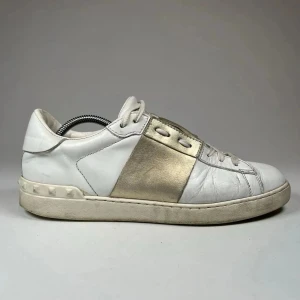 Valentino Garavani Open sneakers vit/guld - Snygga Valentino Garavani Open sneakers i vitt skinn med bred metallic guldpanel över snörningen. Klassisk låg modell med rund tå, platt sula och ikoniska nitar på hälen. Perfekta för dig som vill ha en lyxig och trendig touch till din outfit.