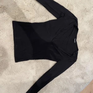 Svart v-ringad topp Gina Tricot  - En svart långärmad topp från Gina Tricot i storlek XS. Toppen har en snygg v-ringning och figurnära passform som framhäver siluetten. Tillverkad i mjuk bomullsmix, perfekt att styla med jeans eller kjol för en clean look.