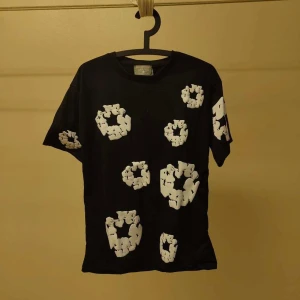 Denim Tears svart - Svart t-shirt från Denim Tears med unika vita blommönster tryckta över hela plagget. Klassisk passform med rund hals och korta ärmar. Tillverkad i mjuk bomull som känns skön mot huden. Perfekt för dig som vill sticka ut med en cool och modern stil.