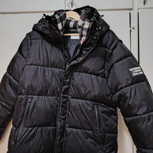 Svart dunjacka Burberry med avtagbara ärmar - Svart dunjacka med avtagbara ärmar så du kan bära den som väst. Jackan har huva, dragkedja, tryckknappar och två fickor med dragkedja. Insidan av huvan har klassiskt rutigt foder i svart, vitt och rött. Perfekt för kalla dagar. Samt höst och vår och sommar alltså kan bli en väst om man vill det. 