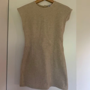 Beige stickad kortklänning från Zara - Superfin beige stickad klänning från Zara i kort modell. Klänningen har en rak passform, markerade axlar och rund halsringning. Perfekt för dig som gillar en enkel men trendig look. Dragkedja i ryggen för enkel påklädning. Storlek S men den är liten i storleken.