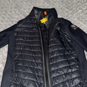 Svart pufferjacka från Parajumpers - Säljer en svart pufferjacka från Parajumpers med quiltad framsida och ärmar i stickad struktur. Jackan har dragkedja, hög krage och en gul detalj vid halsen. Märkespatch på ena ärmen och bröstficka med dragkedja. Perfekt för kyliga dagar.