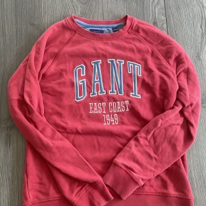 Röd GANT sweatshirt  - Röd sweatshirt från GANT med stort tryck på bröstet där det står 'GANT EAST COAST 1949'. Tröjan har rund hals, långa ärmar och ribbade muddar vid ärmslut och nederkant. Skön och klassisk college-look som passar perfekt till jeans.