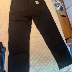 Svarta single knee pants från Carhartt - Svarta cargobyxor från Carhartt med raka ben och flera praktiska fickor, inklusive stora fickor på låren och bakfickor med Carhartt-logga. Byxorna är tillverkade i slitstark bomull och har en klassisk, loose passform.