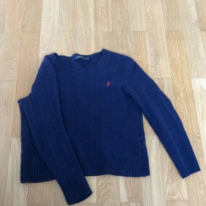 Marinblå kabelstickad tröja Polo Ralph Lauren - Kabelstickad marinblå tröja från Polo Ralph Lauren med klassisk röd broderad logga på bröstet. Tröjan har rund halsringning, ribbade muddar och är långärmad. Inte vanlig storlek i och med krympning i tvätt. Hämtas i Östersund eller skickas på post.