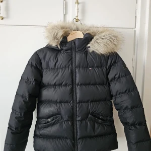 Svart pufferjacka Tommy Hilfiger - Svart pufferjacka från Tommy Hilfiger med huva och snygg fuskpäls runt kanten. Jackan har dragkedja framtill, två fickor med knapp och klassisk quiltad design. Perfekt för kalla dagar och riktigt skön att bära.