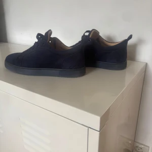 Mörkblå mockasneakers med röd sula - Snygga mörkblå sneakers i mocka från Christian Louboutin med ikonisk röd sula. Skorna har rund tå, snörning och diskret logga på sidan. Perfekta för dig som vill ha en clean men lyxig look med en twist.