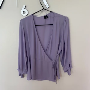 Ljuslila omlottblus från Gina Tricot - Ljuslila blus från Gina Tricot med omlottmodell och knyt i sidan. Långa ärmar med knyt vid handlederna ger en extra detalj. Tunn och flowy känsla, perfekt för vår och sommar. Snygg v-ringning och lätt figursydd passform. Använd fåtal gånger.