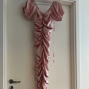 Rosa långklänning med puffärm - Supersnygg långklänning i glansigt rosa satinmaterial med puffiga kortärmar och djup v-ringning. Klänningen har snygg rynkdetalj framtill och snörning i sidan för extra effekt. Funkar för S och M då dennär stretchig.