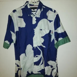 Blommig skjorta Tommy Hilfiger M - Snygg kortärmad skjorta från Tommy Hilfiger i blått med stora vita blommor och gröna detaljer på ärmsluten. Skjortan har klassisk krage, bröstficka med logga och knäppning framtill. Regular fit och perfekt för dig som vill sticka ut. Skjortan är i nyskick, aldrig provat innan! Bra pris
