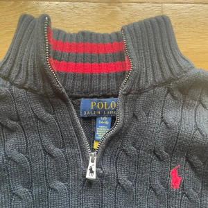 Mörkblå stickad tröja Polo Ralph Lauren - Mörkblå kabelstickad tröja från Polo Ralph Lauren med halv dragkedja och röd broderad logga på bröstet. Hög krage med röda ränder på insidan och långa ärmar. Perfekt för kyliga dagar och snygg till jeans. Sitter mindre än original storleken