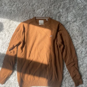 Brun ulltröja från GANT, storlek M - Snygg brun stickad tröja från GANT i superfin lammull. Klassisk rund halsringning med ribbade muddar och diskret broderad logga på bröstet. Perfekt för lager-på-lager och chill höst- eller vinterstil.